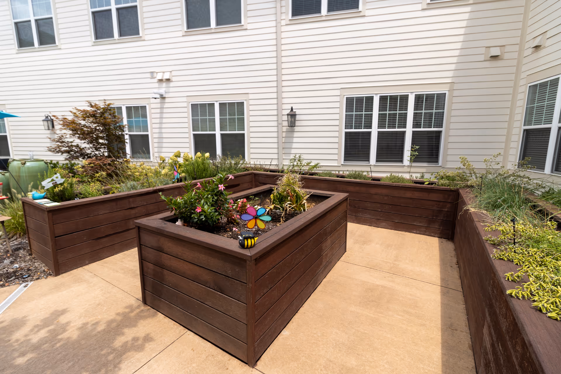 Raised wooden planter boxes with flowers and garden decorations in a courtyard beside a beige multi-story building.