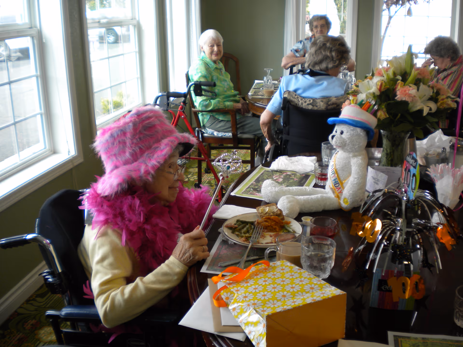 Elderly women sitting around a dining table in a well-lit room with large windows. One woman in the foreground is wearing a bright pink feathered hat and boa, holding a wand, and sitting in a wheelchair. The table is decorated with a birthday-themed teddy bear, flowers, a gift box, and party decorations. Other women are seated around the table, some in wheelchairs, engaging in conversation.