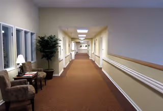 A long, well-lit hallway in a senior living facility with beige walls, brown carpet, handrails along the walls, and a few chairs and a small table with a lamp and plant on the left side.