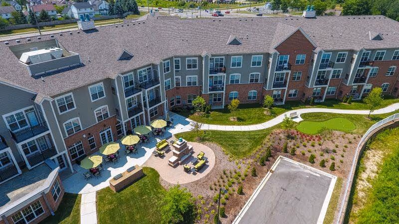 Aerial view of a three-story assisted living building with a landscaped courtyard featuring patio seating with umbrellas, walkways, and a small putting green.