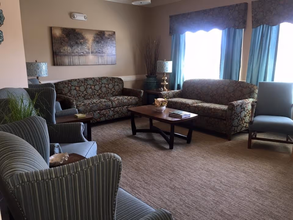 A cozy living room with two floral patterned sofas, two striped armchairs, a wooden coffee table with books and a flower vase, two table lamps with patterned shades, a large painting of trees on the wall, and windows with blue curtains letting in natural light.
