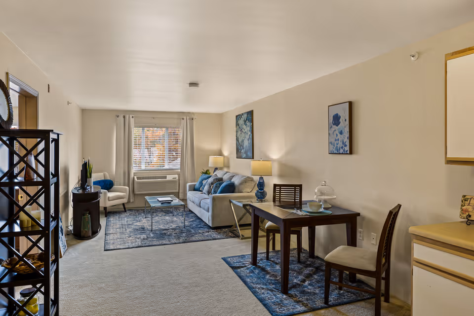 A cozy living and dining area in a senior living facility featuring a beige sofa with blue cushions, a glass coffee table on a blue patterned rug, a small dining table with two chairs on a matching rug, a white armchair near a window with blinds, and decorative wall art. The room has neutral walls and carpeted flooring.
