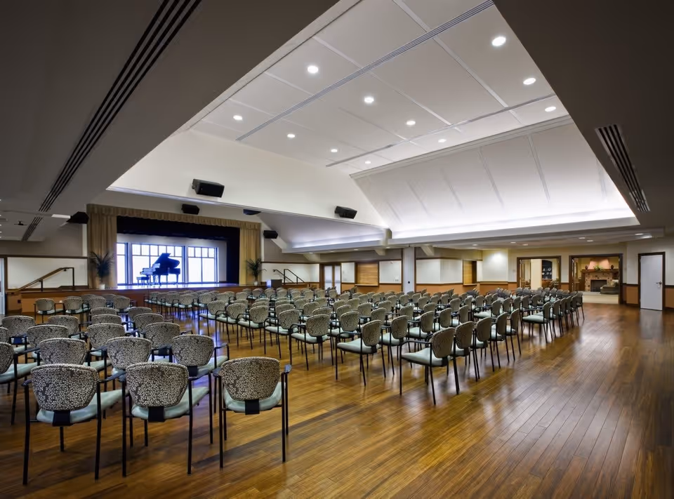 Large auditorium with rows of patterned chairs facing a stage with a grand piano. The room has a high ceiling with recessed lighting and wooden flooring. There are plants on either side of the stage and a cozy sitting area with a fireplace visible through an open doorway to the right.