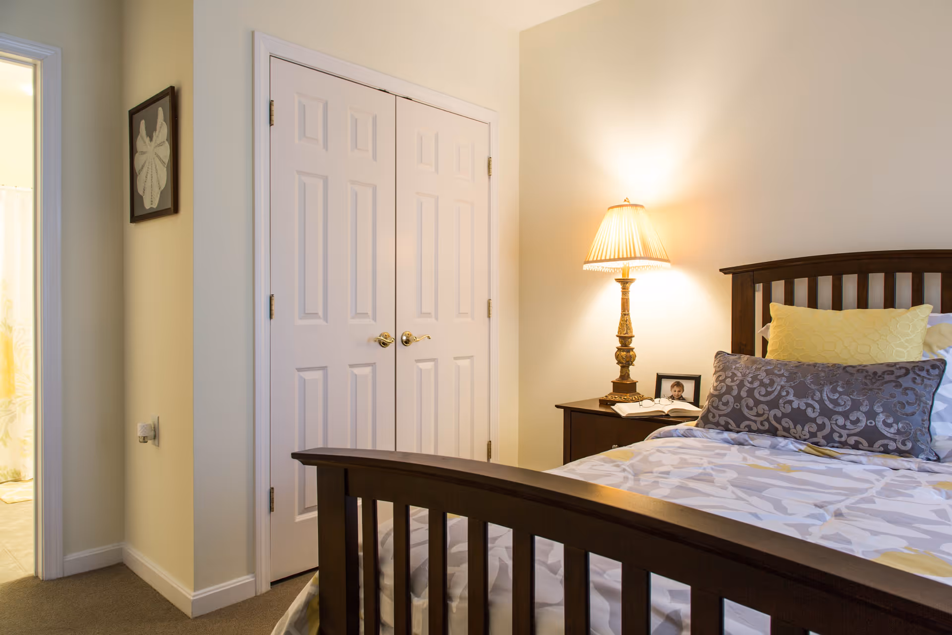 A cozy bedroom with a wooden bed frame, a bed made with patterned bedding and decorative pillows, a nightstand with a lit table lamp, a framed photo, and a book. There is a double-door closet and a glimpse of a bathroom with a shower curtain visible through an open door.