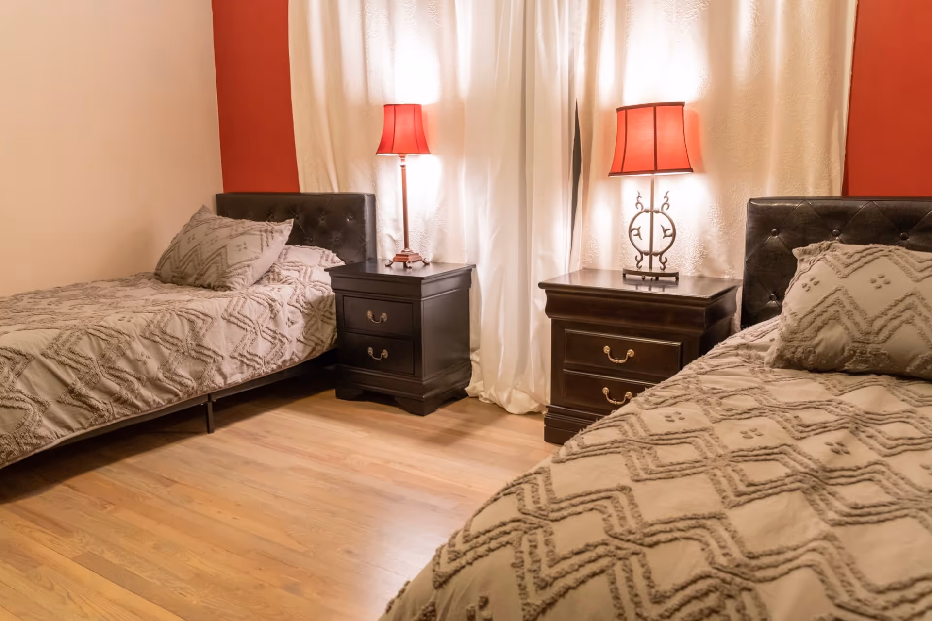 A bedroom with two twin beds featuring tufted dark headboards and beige patterned bedding. Between the beds are two dark wooden nightstands, each with a red lampshade lamp. The room has light wooden flooring and white curtains with a red accent wall behind the beds.