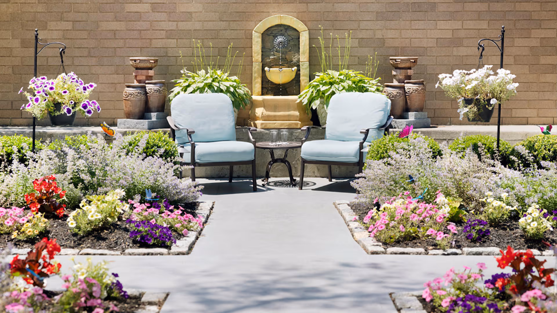 Outdoor seating area with two cushioned chairs and a small glass table between them, surrounded by colorful flower beds and plants. A decorative water fountain is mounted on a brick wall in the background, with potted plants and hanging flower baskets on either side.