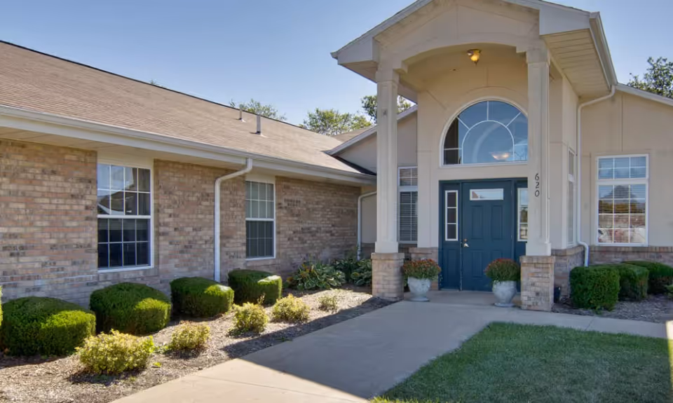 Exterior view of a senior living facility entrance with a covered porch supported by columns, a blue door, brick walls, windows, and neatly trimmed bushes along the walkway.