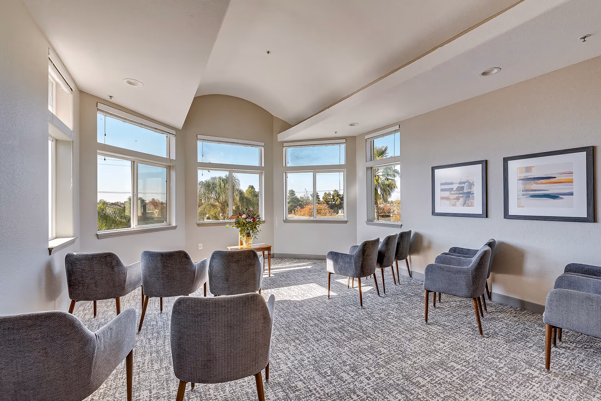 A bright and airy room with large windows letting in natural light, featuring two rows of gray upholstered chairs facing each other, a small wooden table with a vase of flowers near the windows, and two abstract paintings on the wall.