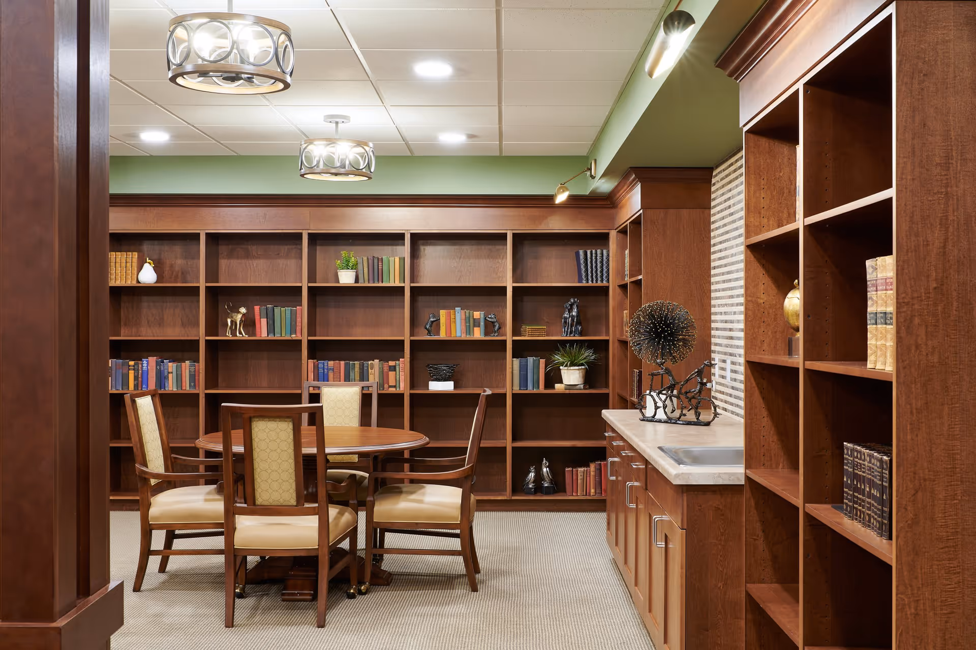 A cozy interior room with wooden bookshelves filled with books and decorative items. In the center, there is a round wooden table surrounded by four cushioned chairs. The room has a beige carpet, green walls, and ceiling lights with circular designs. On the right side, there is a countertop with a small sink and more shelves above and below it.