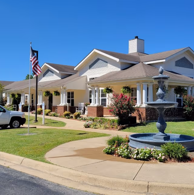 Exterior view of a single-story senior living facility building with white siding and brick accents. The building features a covered porch with white columns, hanging plants, and rocking chairs. In front of the building is a circular fountain surrounded by flowers and greenery. An American flag is displayed on a flagpole near the sidewalk, and a white vehicle is parked nearby under a clear blue sky.