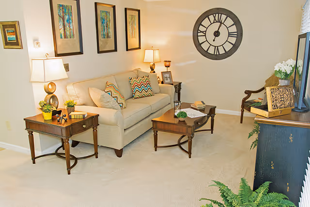 A cozy living room with a beige sofa adorned with patterned and plain cushions, two wooden side tables with lamps and decorative items, a wooden coffee table with a book and small plant, a large wall clock, framed artwork on the wall, a wooden chair, a TV on a stand, and green plants on the floor.