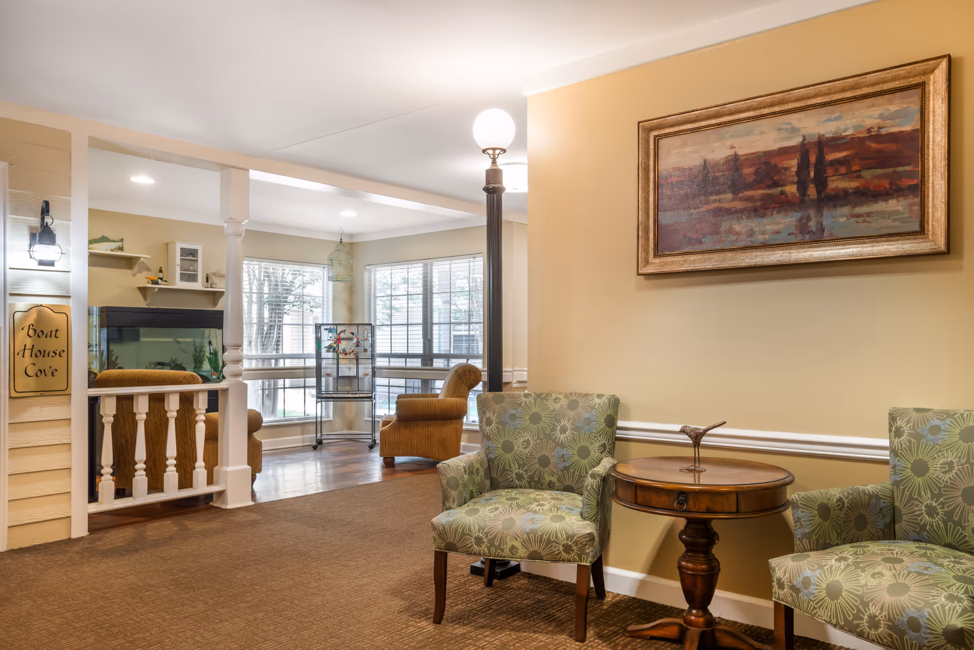 Comfortable interior seating area with two patterned armchairs and a round wooden side table, opening into a bright lounge with large windows and an aquarium.