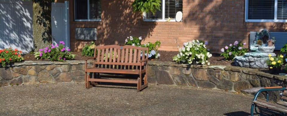 Outdoor seating area with a wooden bench in front of a stone wall planter filled with colorful flowers, next to a brick building with windows and an air conditioning unit.