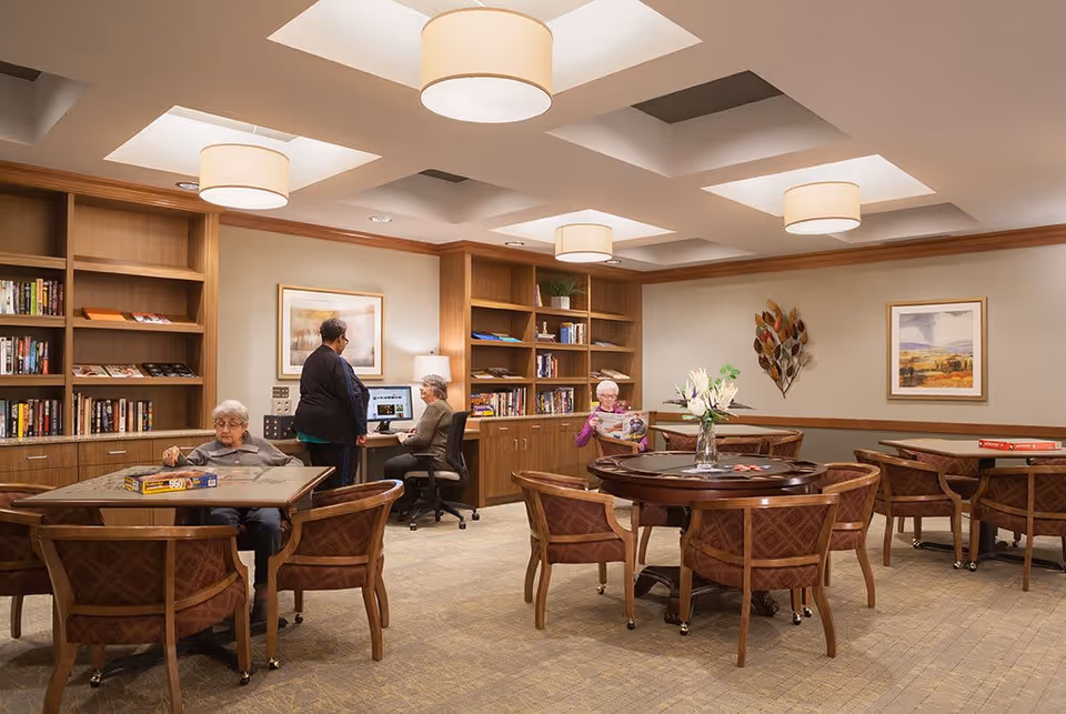 A well-lit common room in a senior living facility with wooden bookshelves, tables, and chairs. Three elderly individuals are present: one is working on a puzzle at a table, another is reading a magazine at a round table, and a third person is standing near a computer desk where another elderly person is seated. The room has beige walls, framed artwork, and decorative lighting fixtures on the ceiling.