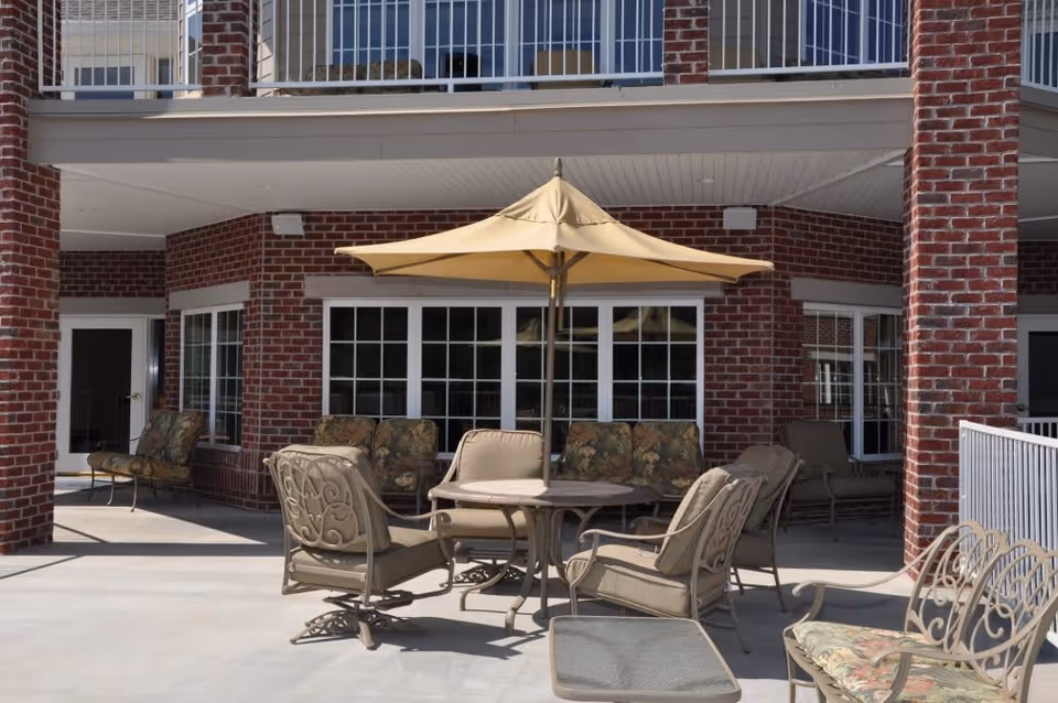 Outdoor patio area with cushioned metal chairs and a round table under a large beige umbrella, set against a brick building with multiple windows and doors.