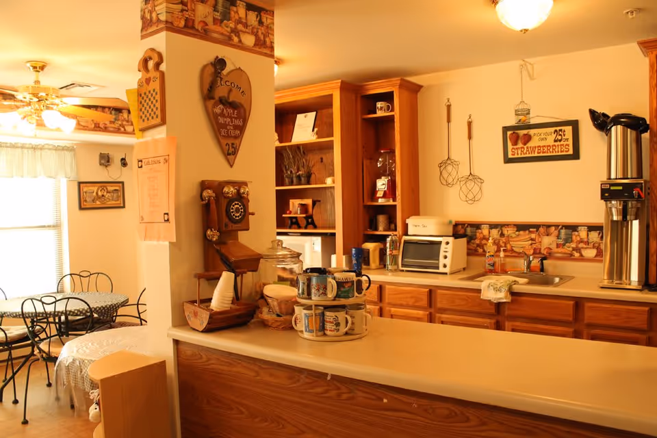 A cozy kitchen and dining area with wooden cabinets, a countertop with stacked mugs, a toaster oven, a coffee dispenser, and a sink. The dining area has a round table with a checkered tablecloth and metal chairs. The walls are decorated with vintage-style signs and kitchen-themed wallpaper border.