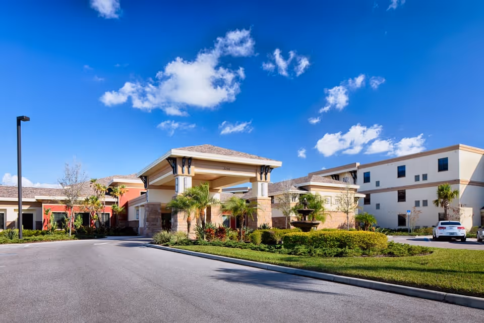 Entrance drive and porte-cochere of The Fountains of Hope with landscaped fountain, shrubs, and a clear blue sky.