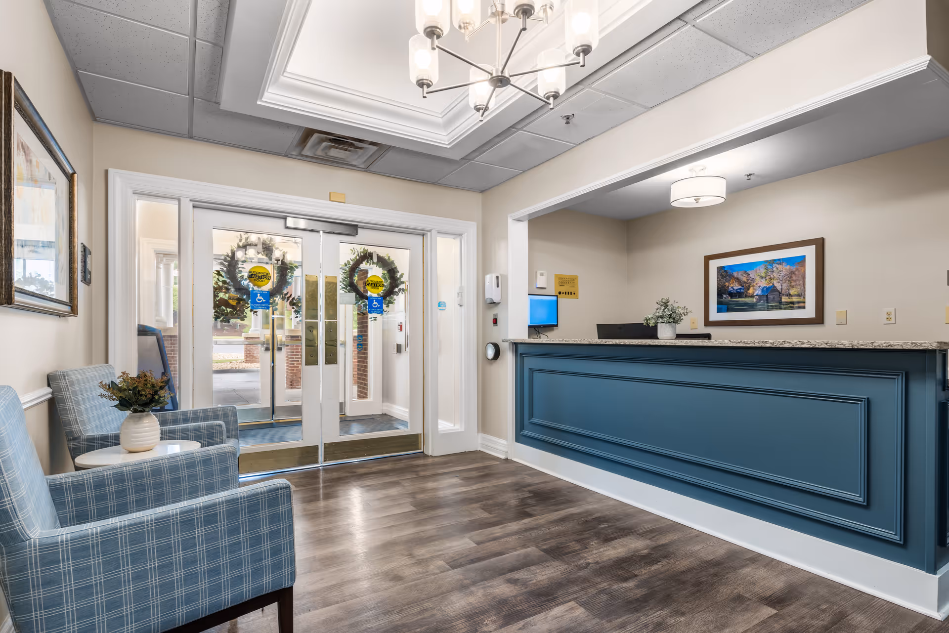 Reception area of TerraBella Pheasant Ridge featuring a blue front desk with a granite countertop, two blue plaid armchairs with a small white table and a vase with flowers, glass double doors decorated with wreaths, and a framed landscape photograph on the wall behind the desk.