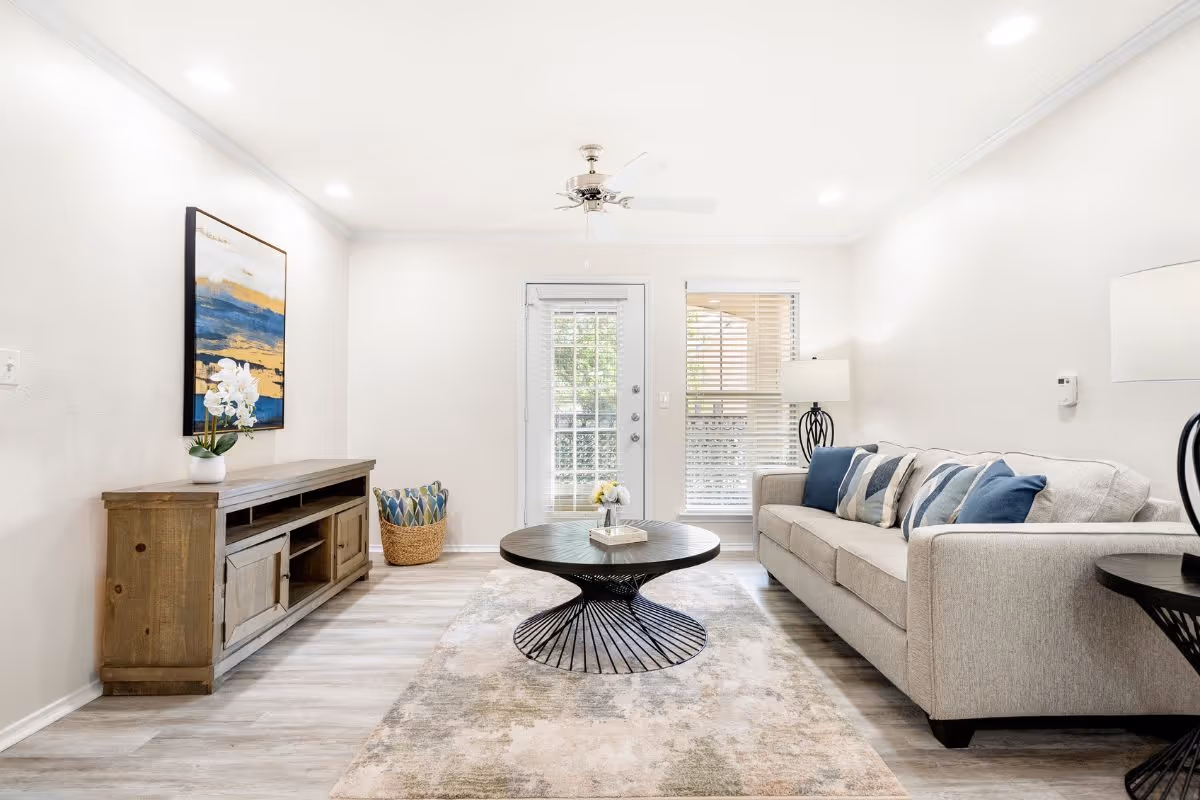 Bright living room with a light-gray sofa, round black coffee table, wooden console, and a glass door with blinds.