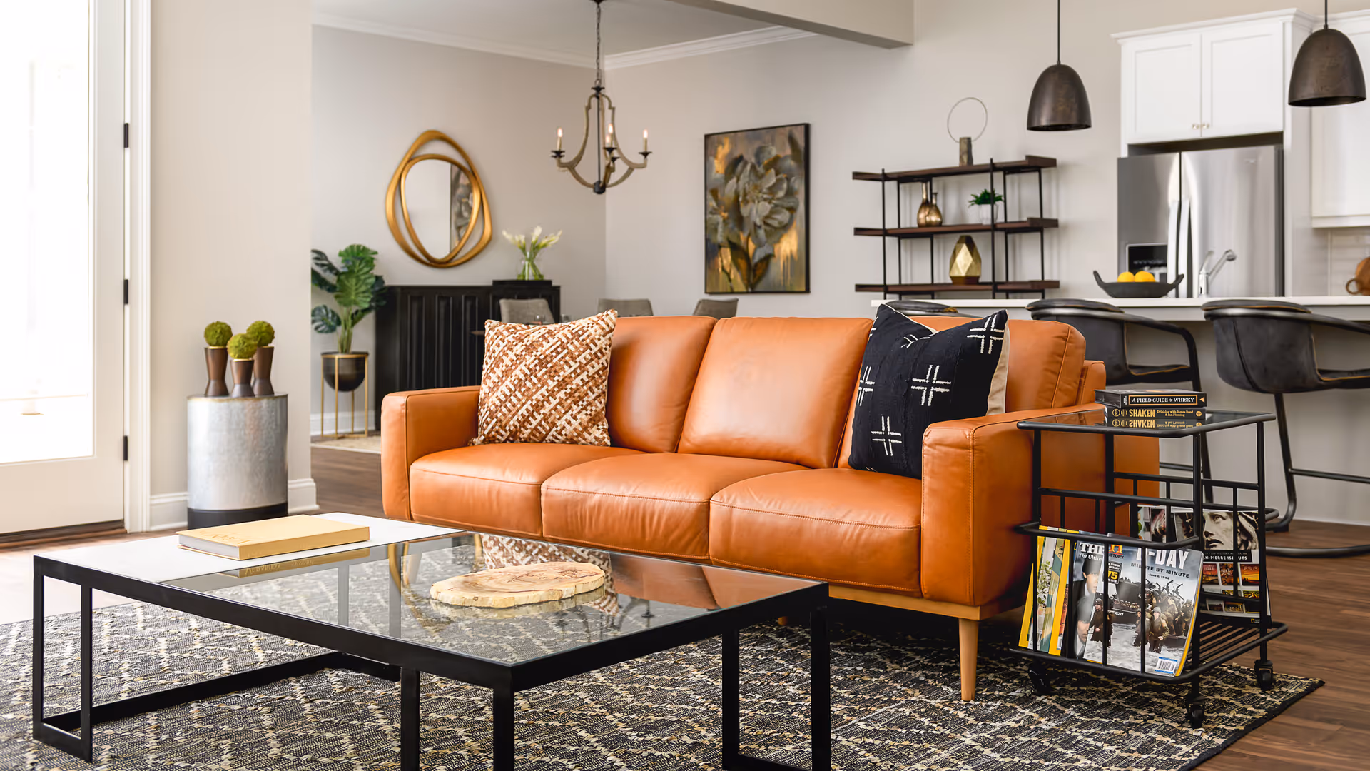 Modern living room with a tan leather sofa, glass coffee table on a patterned rug, and an open kitchen in the background.