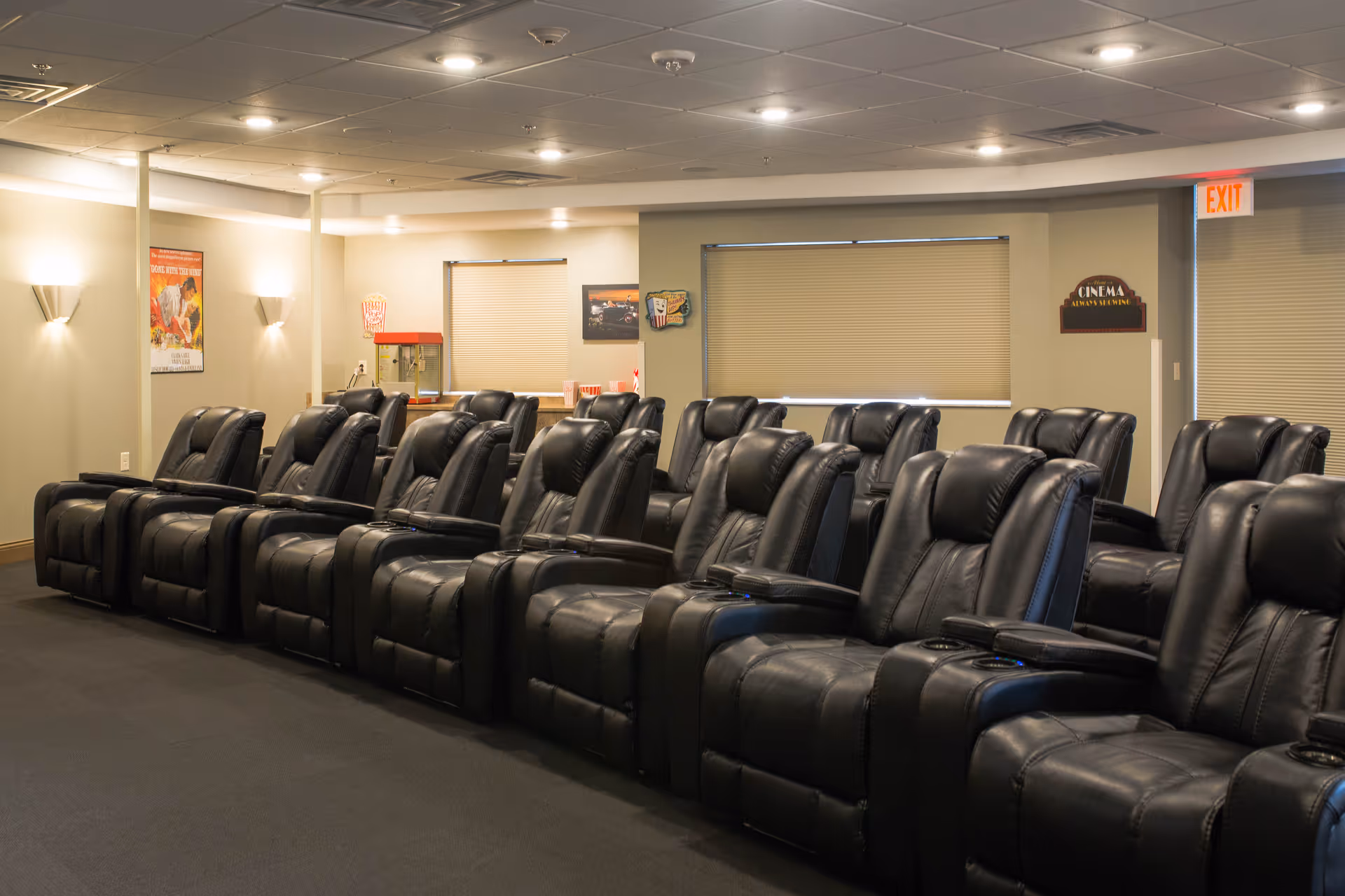 A cozy movie theater room with two rows of black leather recliner chairs facing a screen. The room has beige walls with wall-mounted lights, a popcorn machine in the back corner, movie posters on the walls, and a sign that reads 'Cinema Always Showing'.