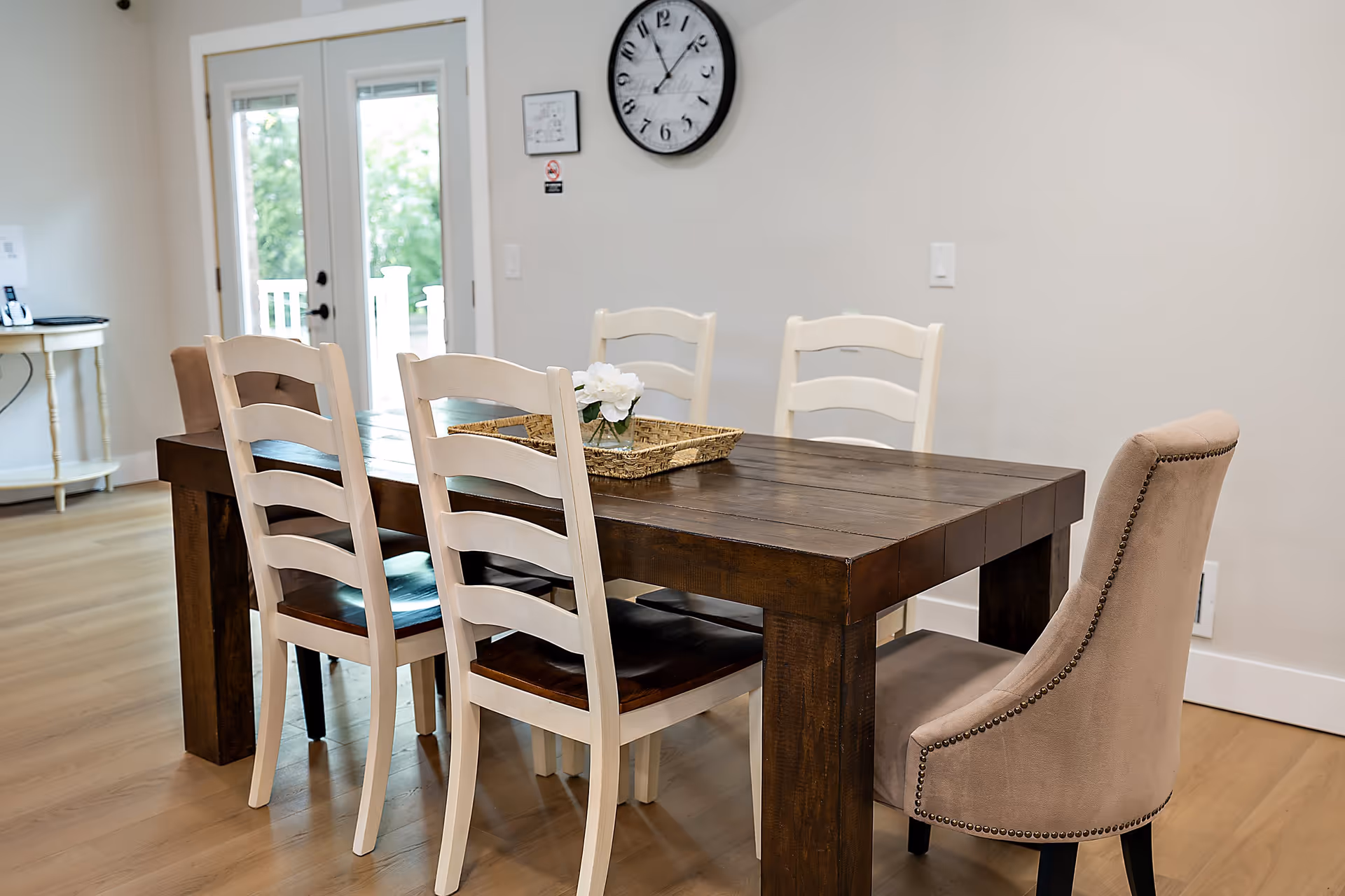 Bright dining area with a dark wooden table surrounded by white ladder-back chairs and a beige upholstered chair, a woven tray with flowers on the table, a wall clock, and French doors in the background.