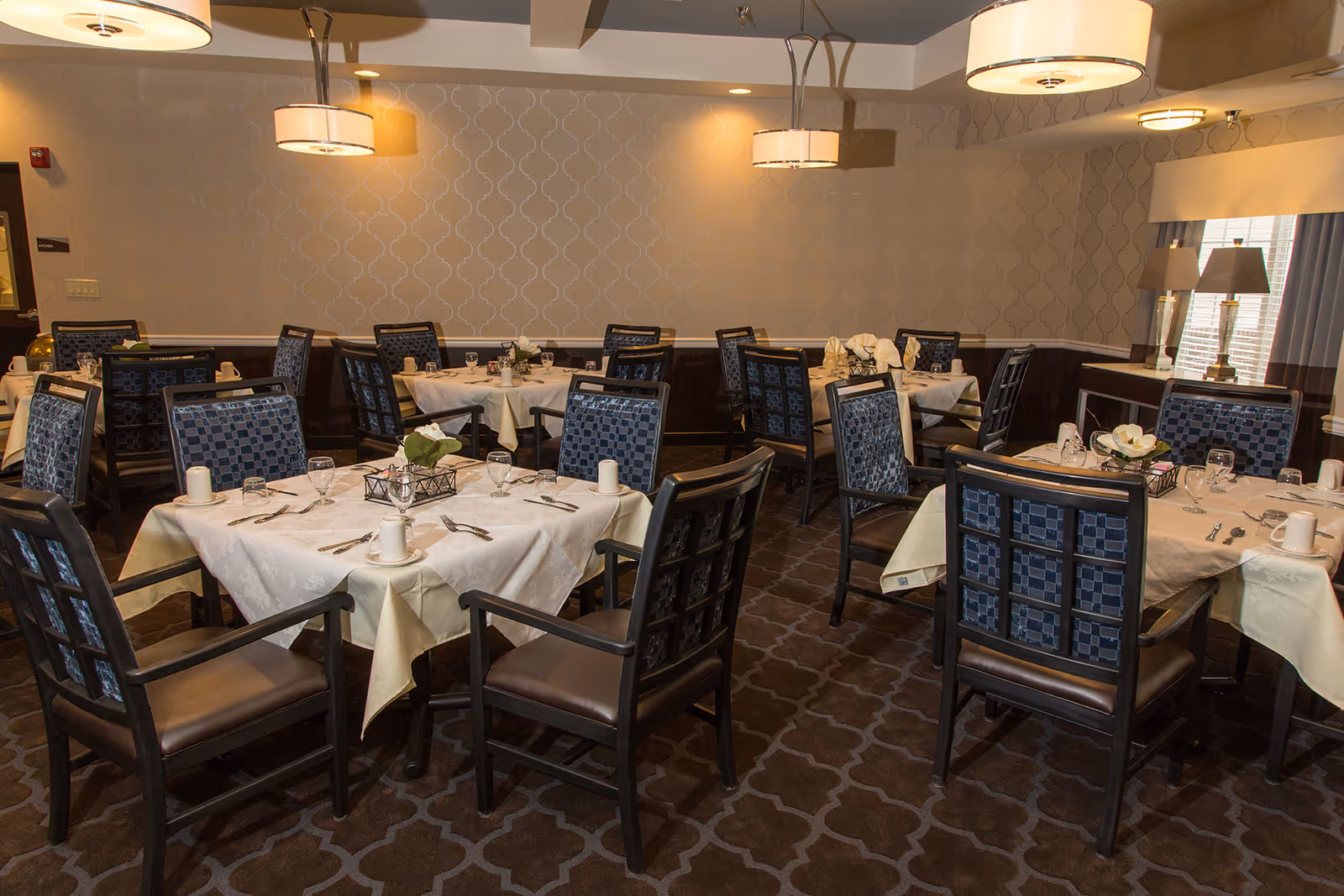 A dining room in a senior living facility with several tables covered in white tablecloths, each set with glasses, cups, silverware, and small floral centerpieces. The room has patterned wallpaper, carpeted floors, and modern hanging light fixtures. There are upholstered chairs with dark wooden frames around each table.
