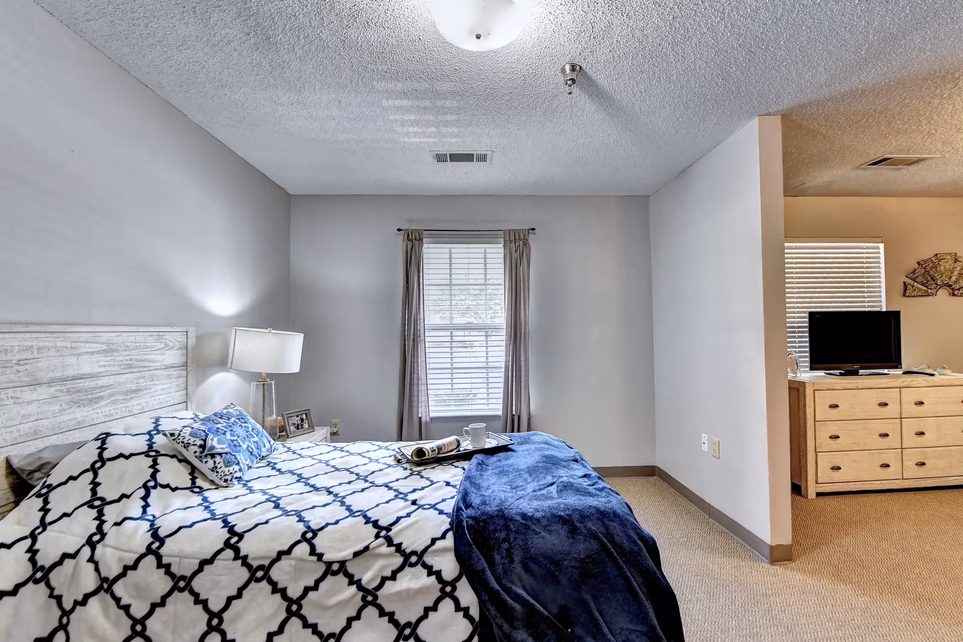 A bedroom with a bed covered in a white and navy blue patterned comforter and a navy blue throw blanket. There is a nightstand with a lamp and picture frames next to the bed. A window with beige curtains is centered on the wall behind the bed. To the right, there is a dresser with a TV on top and a window with blinds behind it. The walls are light-colored and the carpet is beige.