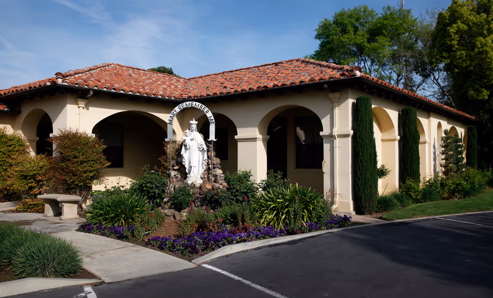 Exterior view of a single-story building with beige walls and a red tile roof. In front of the building is a garden with various green plants and purple flowers, centered around a white statue of Mary with an arch above it that reads 'MARY REMEMBER ME'. There is a concrete bench on the left side and a paved driveway in the foreground.