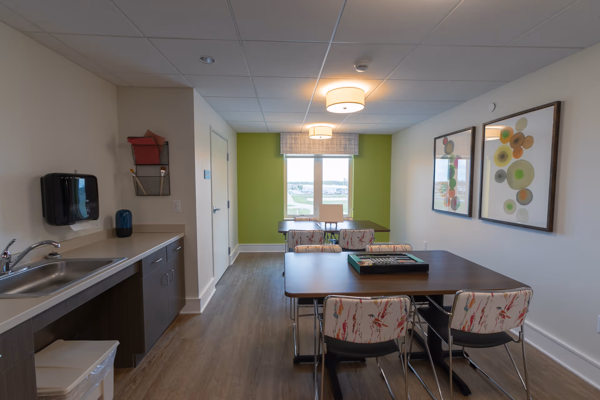A well-lit room with a kitchenette on the left side featuring a sink, countertop, and cabinets. In the center and right side of the room, there are two tables with chairs around them, one table closer to the window and the other in the foreground with a board game on it. The back wall is painted green around the window, and two framed abstract artworks hang on the right wall. The floor is wood, and the ceiling has recessed lighting and two ceiling lamps.