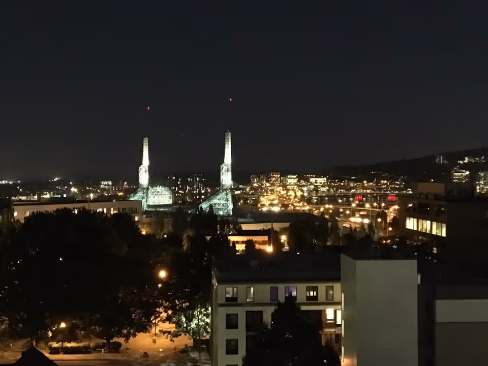 Nighttime cityscape view featuring illuminated buildings and two tall, lit towers with red lights on top, surrounded by other city lights and structures.
