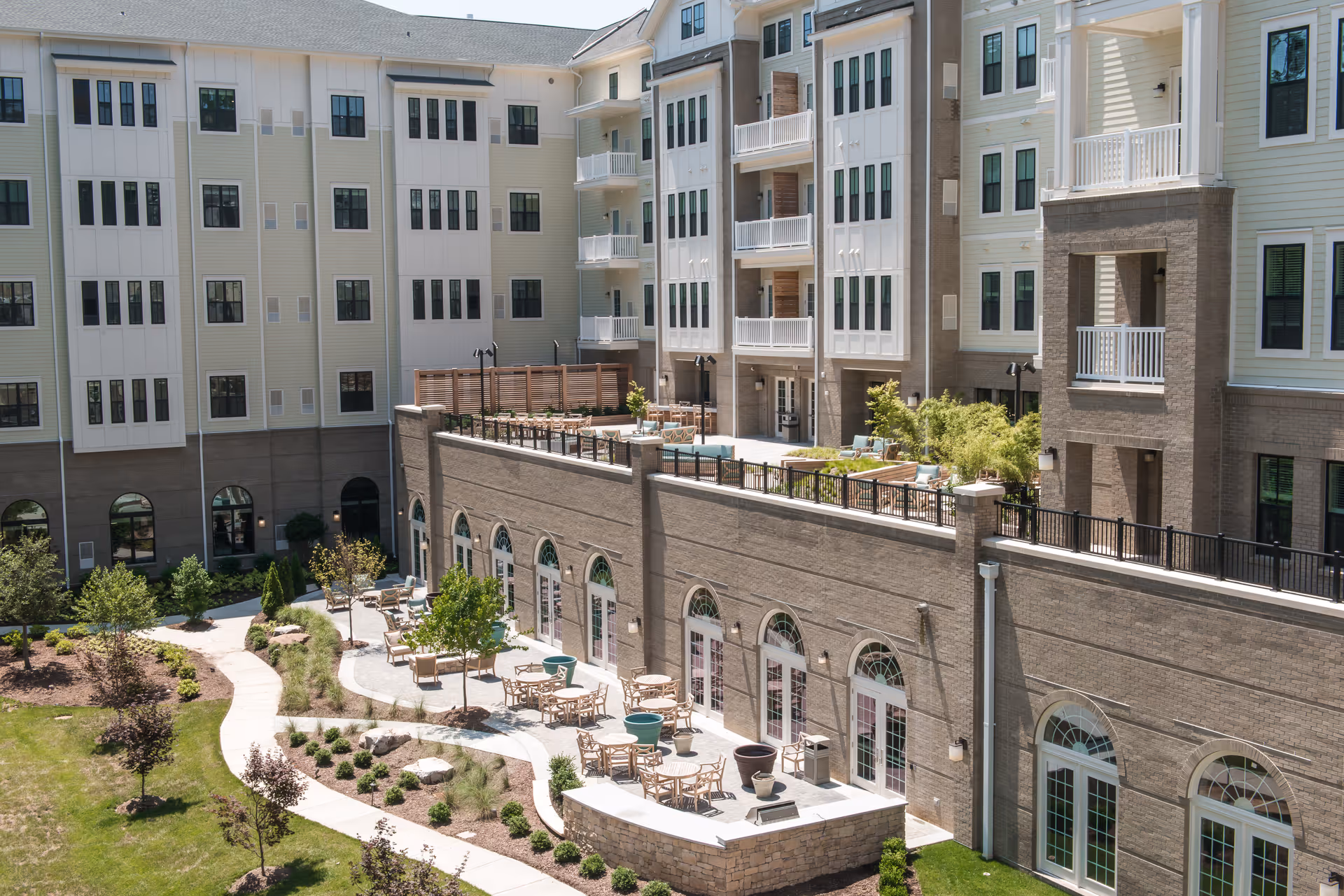 Outdoor patio area of a senior living facility with multiple tables and chairs arranged along a curved walkway, surrounded by landscaped greenery and trees. The multi-story building features balconies and large windows overlooking the patio.