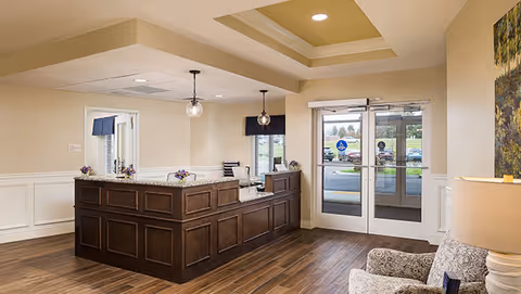 Reception area with a dark wood front desk, two hanging pendant lights, a seating area with patterned armchairs, a lamp, and double glass doors leading outside. The floor is wood, and the walls are painted beige with white wainscoting.