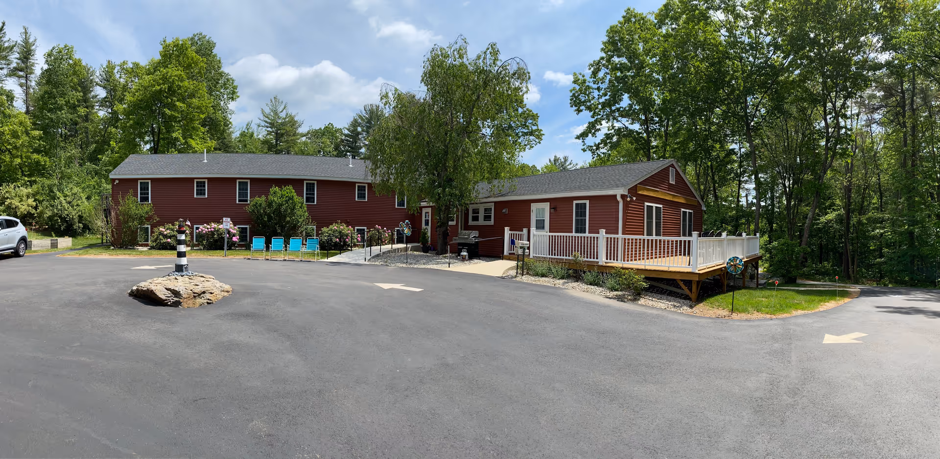 Exterior view of a red building at Assisted Living at Pine Hill Retirement Home surrounded by trees and greenery. There is a paved driveway with a small rock island featuring a black and white striped lighthouse decoration. Several blue chairs are lined up near the building, and a wooden deck with white railings extends from the right side of the building.
