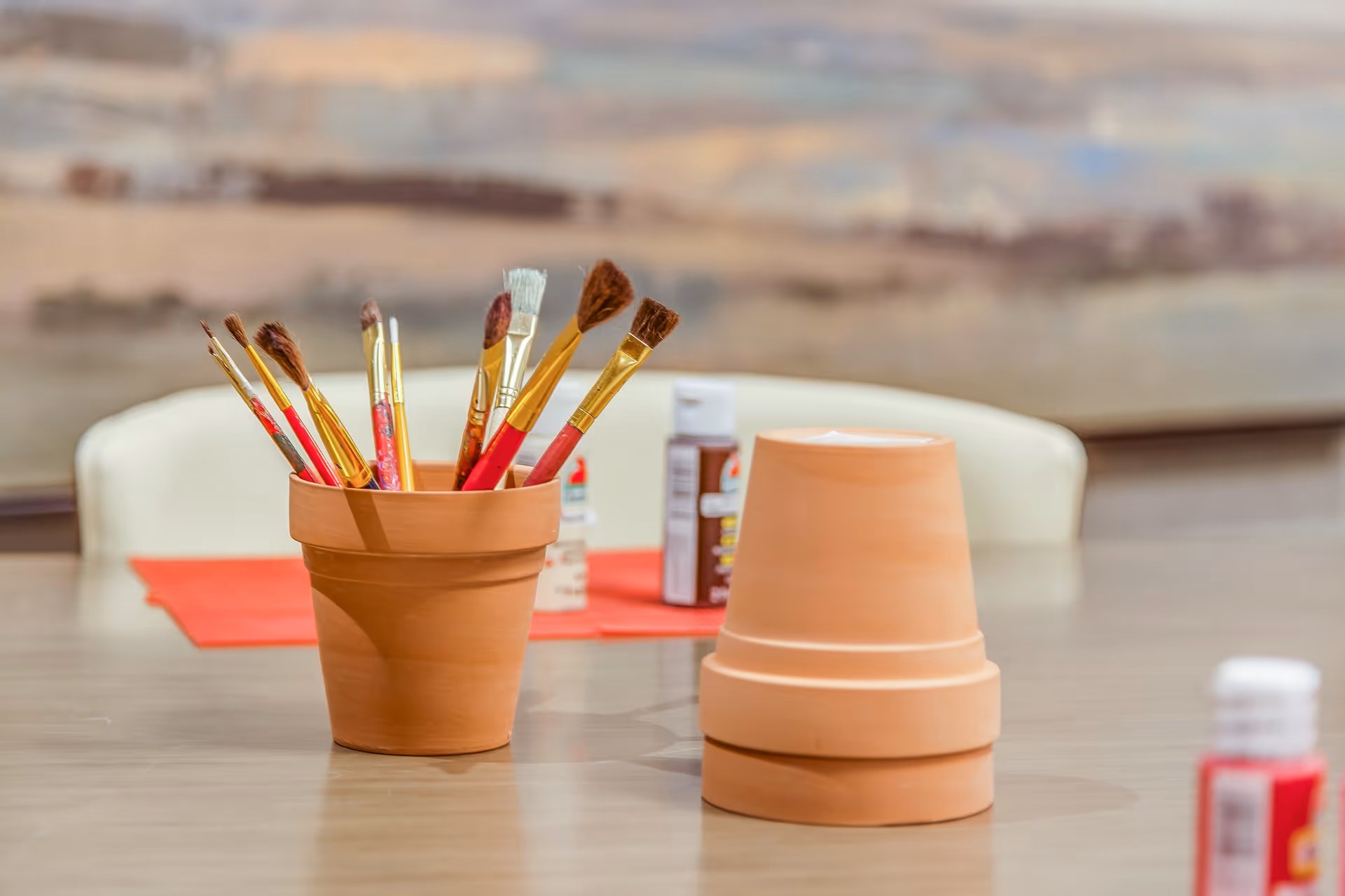 Paintbrushes in a terracotta pot and stacked clay pots on a table with paint bottles in the background.