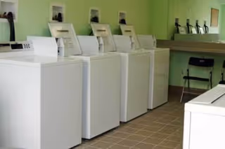 Laundry room with five white top-loading washing machines lined up against a light green wall. There is a folding counter with a black folding chair underneath it on the right side of the room. The floor is tiled with light brown tiles.