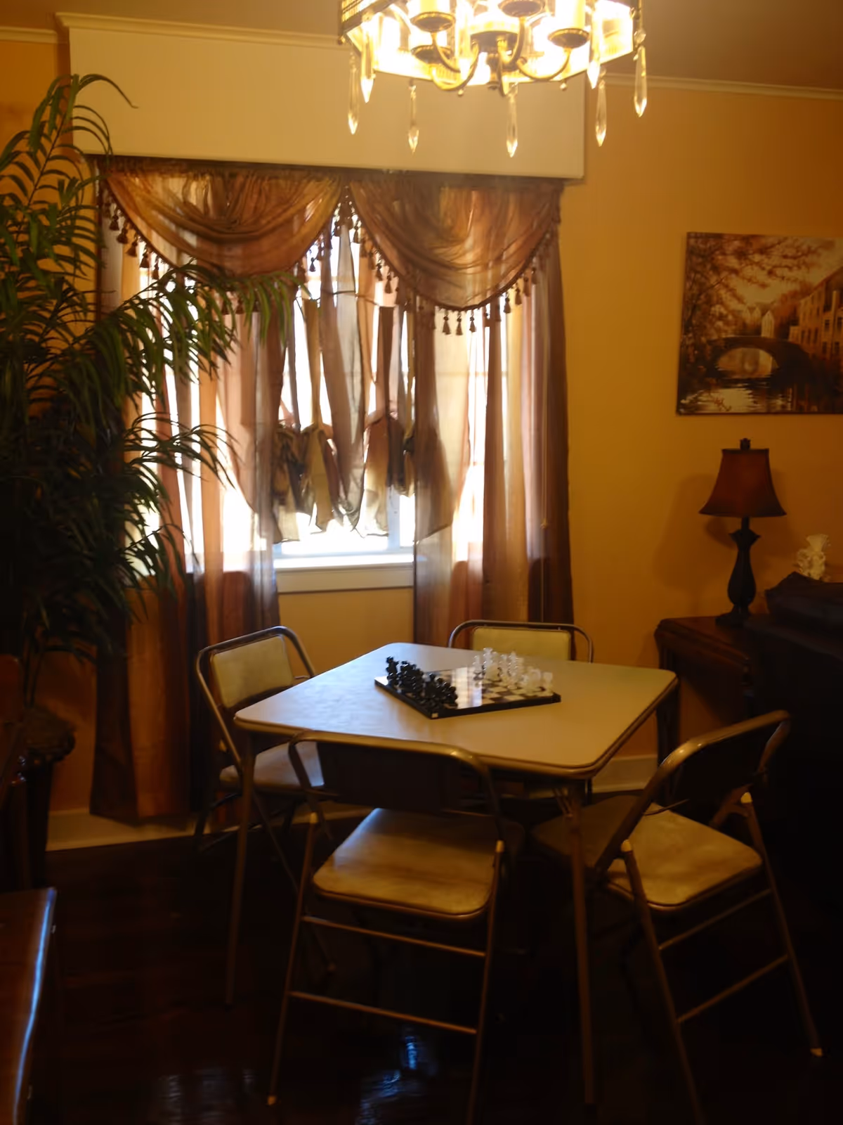 A cozy room with a square table in the center surrounded by four chairs. On the table is a chessboard with chess pieces set up. Behind the table is a window with sheer brown curtains allowing light to filter through. To the right, there is a wooden side table with a lamp and a framed picture on the wall above it. A large green plant is visible on the left side of the image. A chandelier hangs from the ceiling above the table.