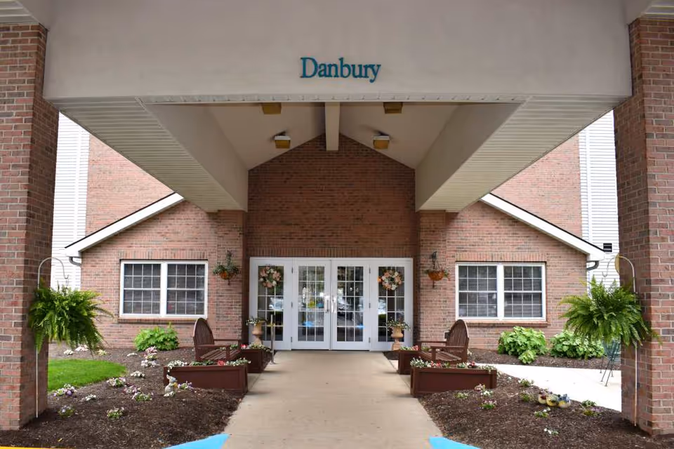 Entrance of a brick building with a covered walkway. The building has double glass doors decorated with floral wreaths. There are two wooden benches on either side of the walkway, surrounded by flower beds and hanging green plants. The word 'Danbury' is displayed above the entrance.