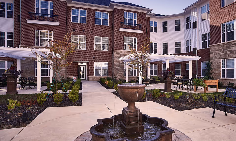 Courtyard in front of a multi-story brick senior living building with a central fountain, pergolas, seating areas, and landscaped walkways.