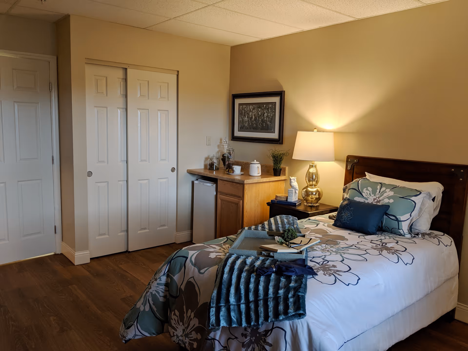 A cozy bedroom with a single bed featuring floral bedding and a blue throw blanket. There is a wooden nightstand with a gold lamp, a small owl figurine, and books. A small countertop area with a mini fridge, jars, a teapot, and a cup is visible against the wall. The room has wooden flooring, beige walls, and a closet with sliding doors.