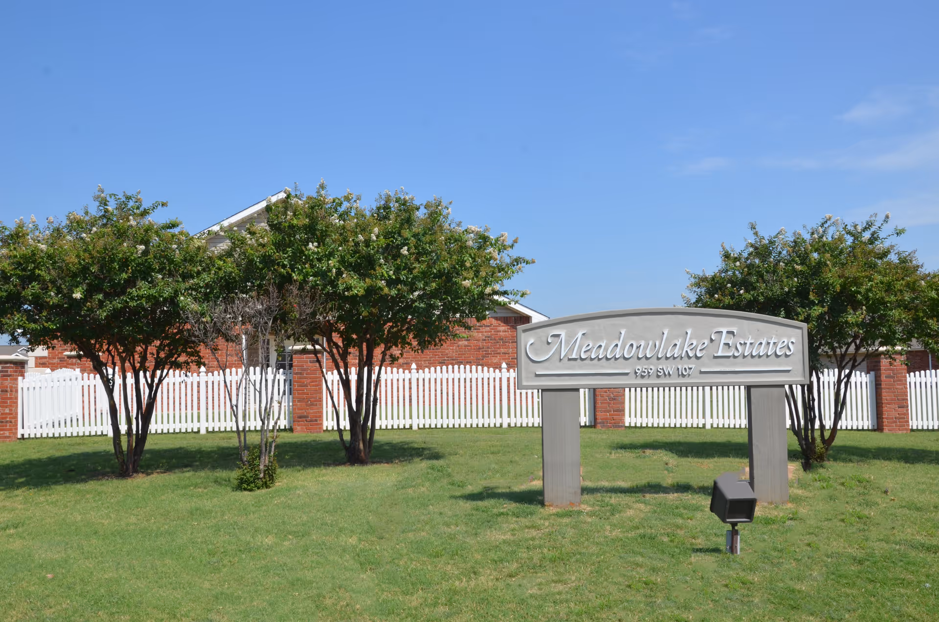 A lawn with trees and a silver 'Meadowlake Estates' sign in front of a white picket fence and brick building under a clear blue sky.
