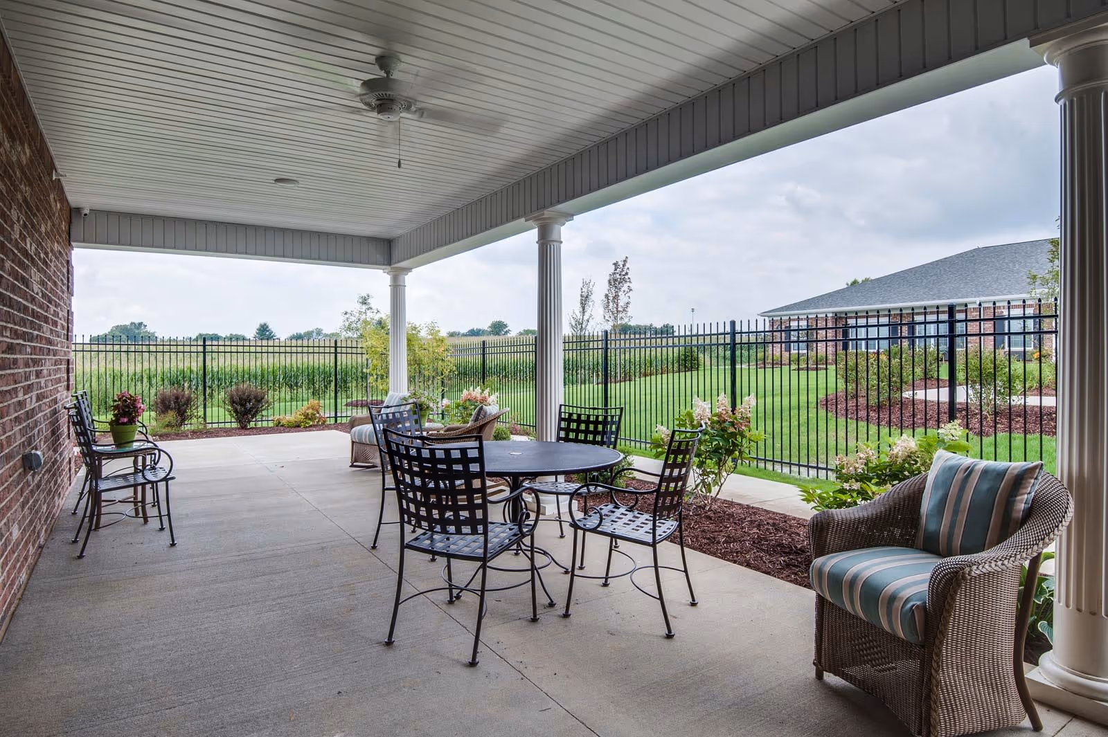 Covered outdoor patio area with metal table and chairs, a wicker armchair with striped cushion, and a ceiling fan. The patio overlooks a fenced grassy area with shrubs and a building in the background under a cloudy sky.