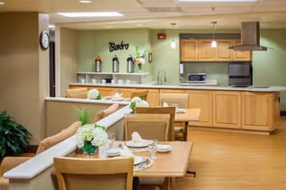 A clean and well-lit dining area with wooden tables and chairs, each table set with plates, glasses, and white floral centerpieces. In the background, there is a kitchen area with wooden cabinets, a microwave, an oven, and a range hood. The walls are painted light green, and the word 'Bistro' is displayed on the back wall above the counter.