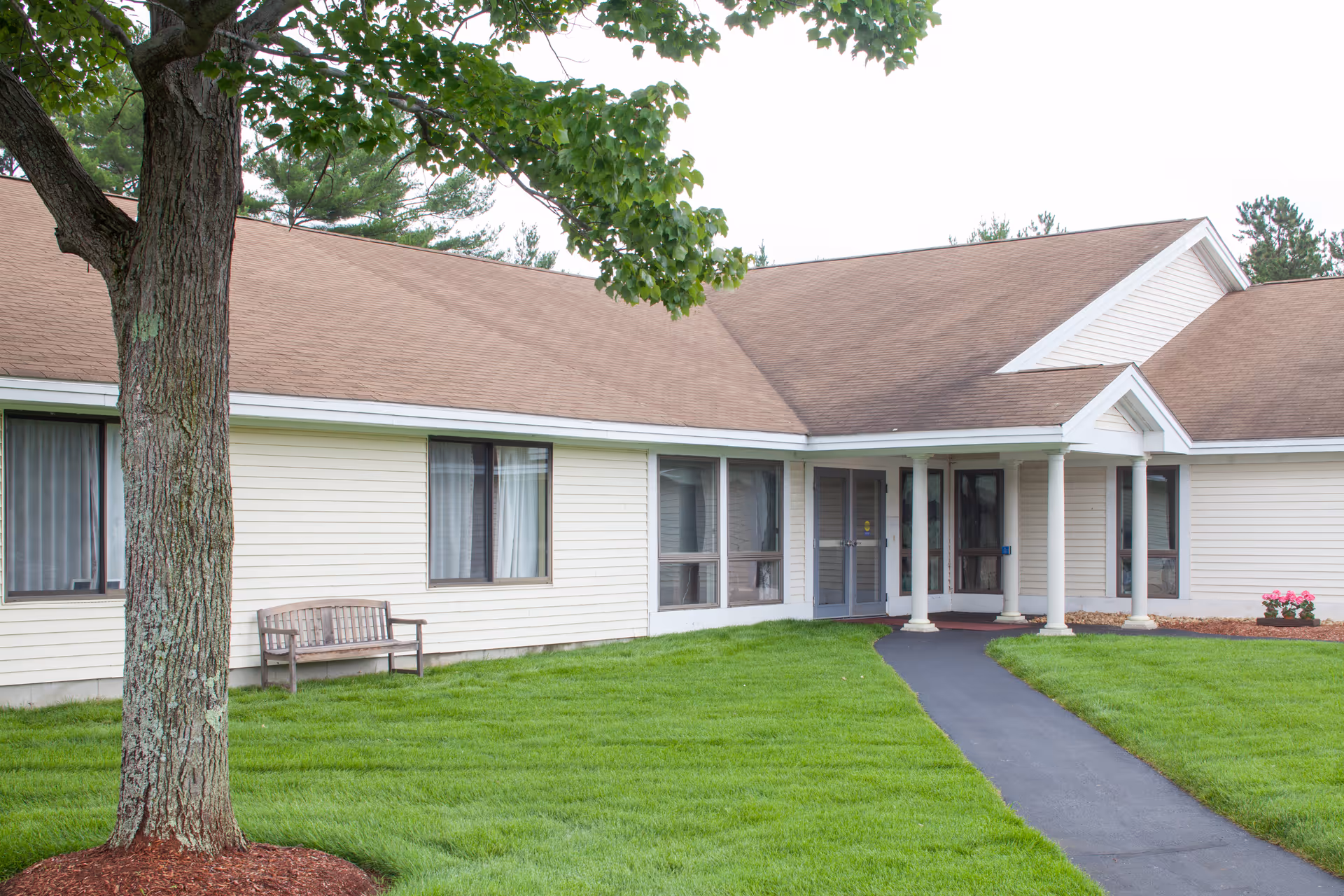Exterior view of a single-story building with beige siding and a brown roof. There is a paved walkway leading to a covered entrance supported by white columns. A tree and a wooden bench are visible on the well-maintained green lawn in front of the building.