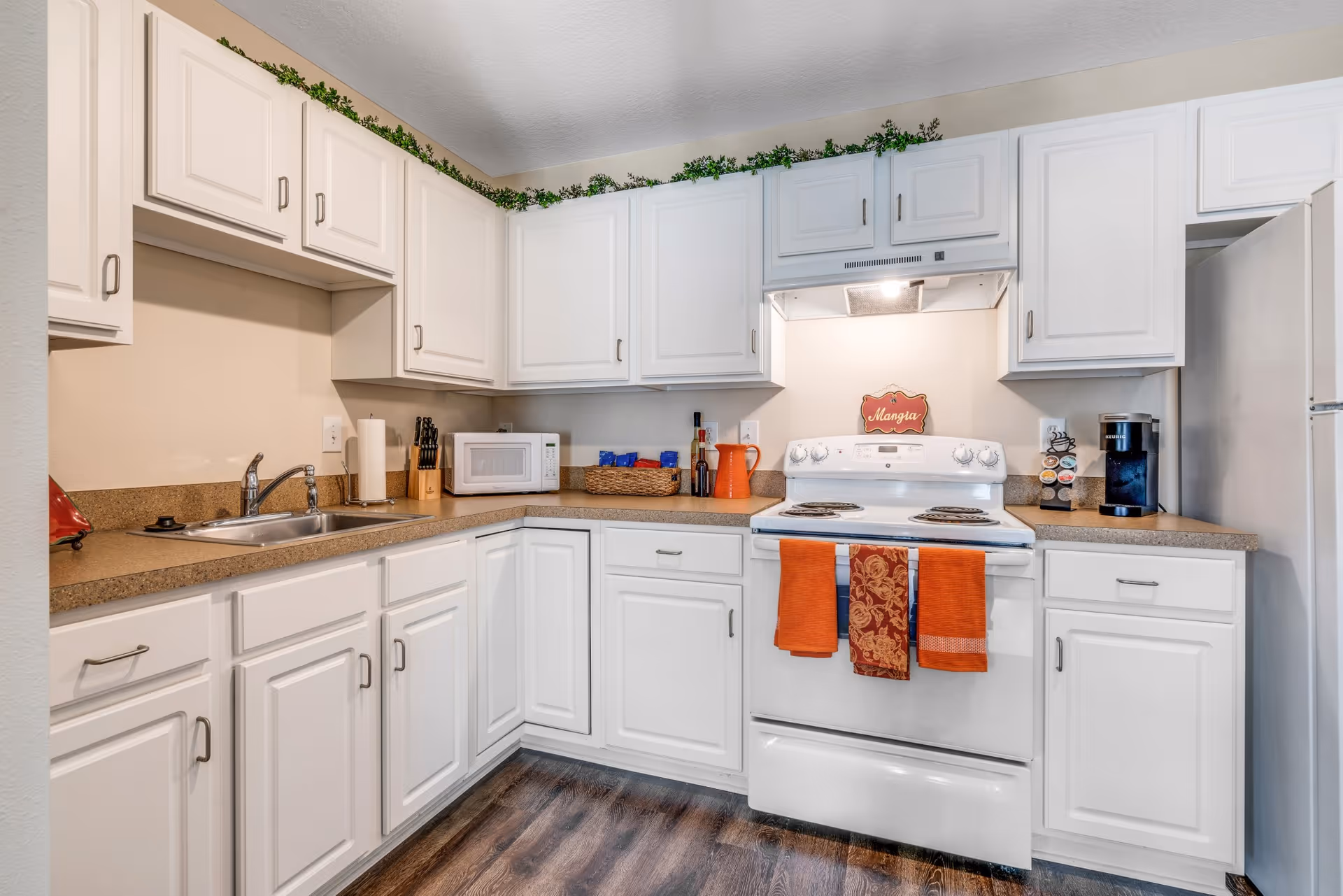 A clean and well-organized kitchen with white cabinets and brown countertops. The kitchen features a white stove with three orange towels hanging on the handle, a microwave, a coffee maker with coffee pods, a sink, and various kitchen utensils. Greenery decorates the top of the cabinets, and the floor has a wood-like finish.