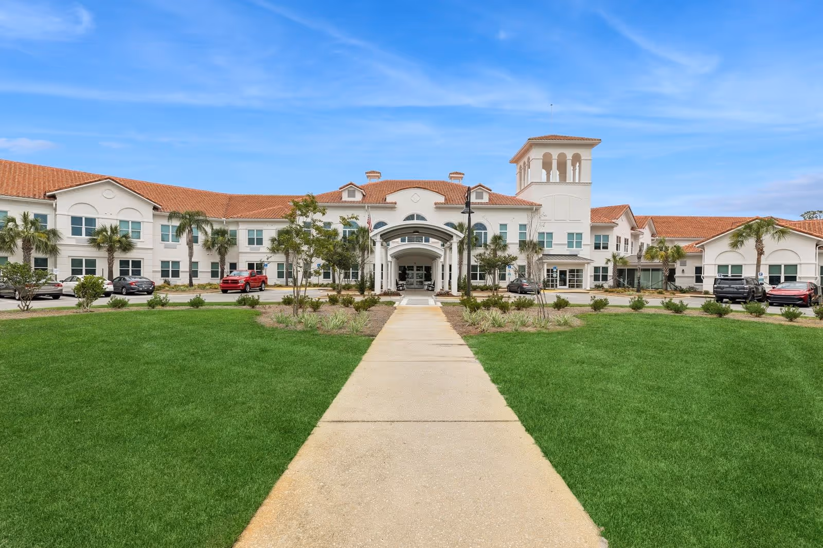 Front exterior view of Somerby Santa Rosa Beach facility with a wide concrete walkway leading to the main entrance, surrounded by green lawns, palm trees, and parked cars under a blue sky.