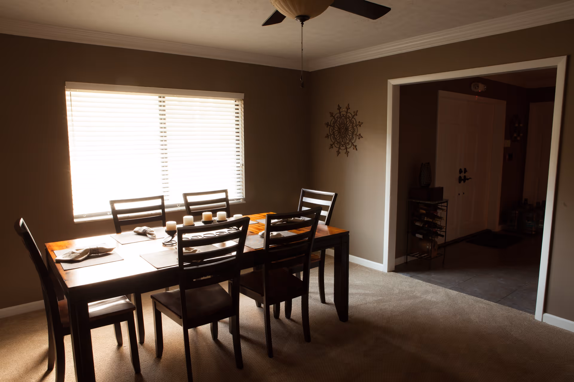 A dining room with a wooden table and six chairs. The table is set with placemats, napkins, and a centerpiece of candles. There is a large window with blinds letting in natural light, a ceiling fan above, and a decorative wall piece on the right wall. An open doorway leads to another room with a white door and some furniture.