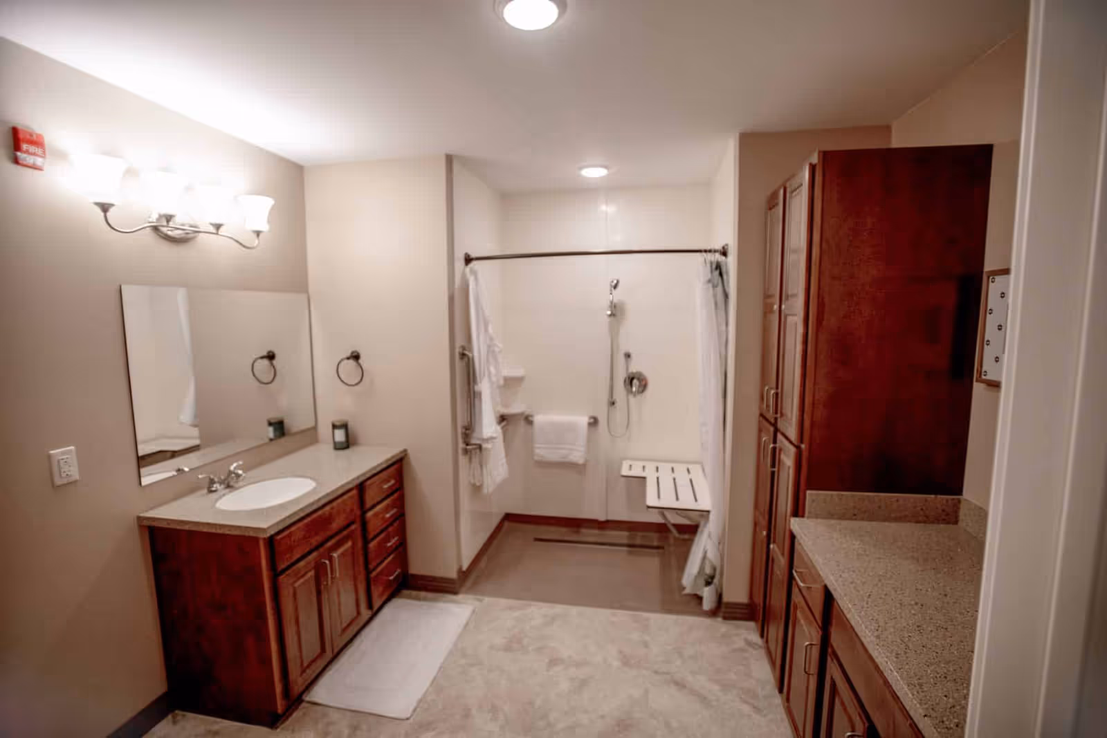A spacious bathroom with beige walls and flooring. On the left side, there is a wooden vanity with a sink, a large mirror, and three light fixtures above it. In the center, there is a walk-in shower with a white curtain, a foldable shower seat, and grab bars. On the right side, there are tall wooden cabinets and a countertop. The bathroom is well-lit with ceiling lights.