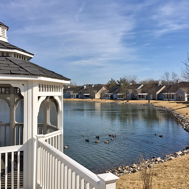 View of a peaceful pond with ducks swimming, surrounded by a row of single-story houses and a white gazebo with a railing in the foreground under a partly cloudy blue sky.