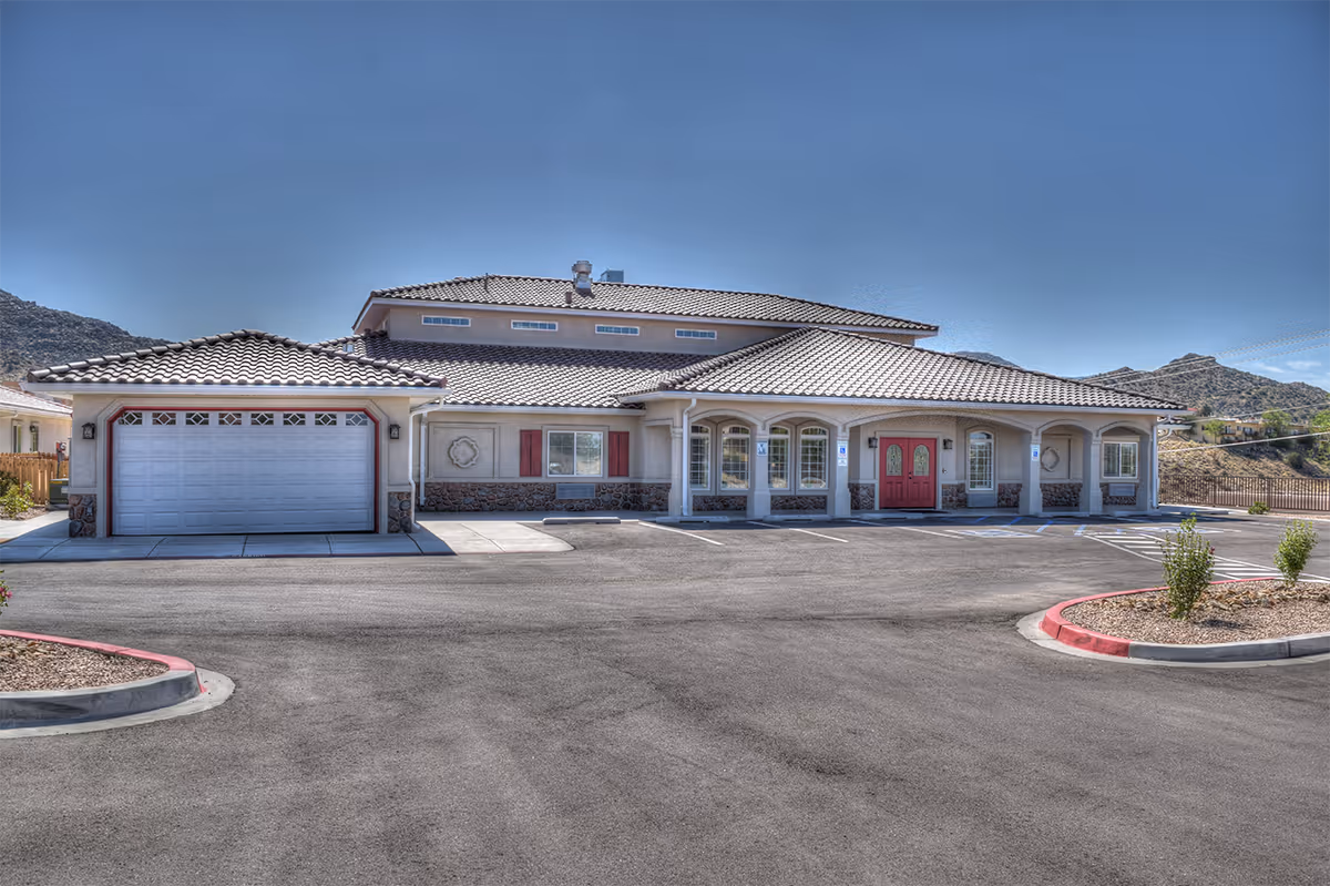 Exterior front view of a single-story building with a tiled roof, red double front doors, multiple windows, and a garage door on the left side. The building is surrounded by a paved parking lot with marked parking spaces and landscaped areas with small plants. Mountains are visible in the background under a clear blue sky.